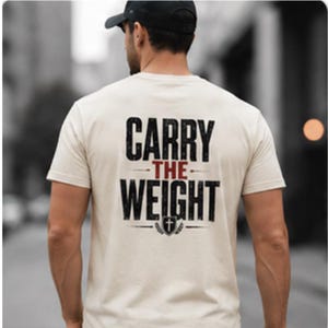 Könnte beinhalten: Ein cremefarbenes Kurzarm-T-Shirt mit schwarzem und rotem Text auf der Rückseite, der CARRY THE WEIGHT lautet, sowie einem kleinen Schildsymbol mit einem Kreuzzeichen unter dem Text. Das T-Shirt wird mit einer schwarzen Baseballkappe getragen.