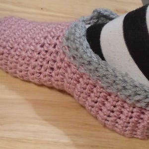 May include: A pink crocheted slipper with a grey trim. The slipper is made of a soft, textured yarn and appears to be handmade. The slipper is on a wooden surface.