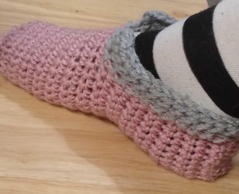 Pu&ograve; includere: Una pantofola rosa all'uncinetto con un bordo grigio. La pantofola &egrave; realizzata con un filato morbido e strutturato e sembra fatta a mano. La pantofola &egrave; su una superficie di legno.