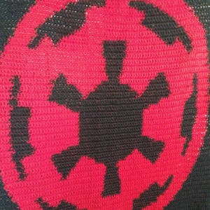 May include: A black and red crocheted blanket featuring a central red Imperial symbol. The symbol is a stylized circular design with a black center and black shapes around the perimeter. The blanket is made of yarn.