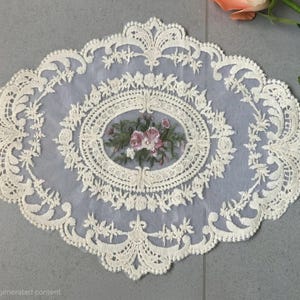 May include: An oval-shaped cream-colored lace doily featuring an intricate embroidered floral motif in the center with pink and green threads. The lace border displays a detailed scalloped edge with symmetrical leaf and vine patterns throughout.