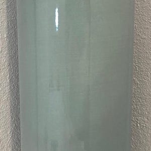 May include: A tall, cylindrical vase in a muted teal color. The vase has a glossy finish and a slightly rounded base. The brand name "Gardenhouse" is visible near the base. The vase is suitable for home decor.