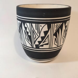 May include: Black and white ceramic pot with a geometric design. The pot has a wide, cylindrical shape with a black exterior and a white interior. The design features repeating geometric shapes in white against the black background, with a white band at the top and bottom.