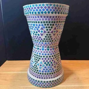 May include: A decorative mosaic vase with a unique hourglass shape. The vase is adorned with a colorful mosaic pattern of blue, purple, and green tiles. The vase is approximately 12 inches tall.