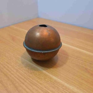 May include: A copper-colored, spherical object with a dark opening at the top. A dark metal band encircles the sphere's midsection. The object sits on a wooden surface.