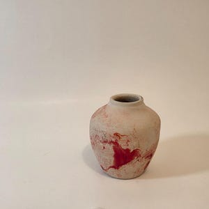 May include: A small, round, ceramic vase with a wide opening. The vase is primarily a light beige color, with red, marbled patterns throughout. The vase has a textured, matte finish and is suitable for displaying flowers or as a decorative accent.