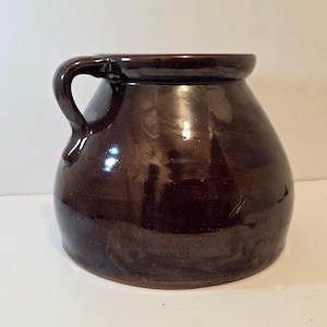 May include: A dark brown, glazed ceramic jug with a rounded body and a curved handle. The jug has a wide opening and a slightly flared rim. The surface has a glossy finish, reflecting light. This antique-style jug is suitable for kitchen or home decor.