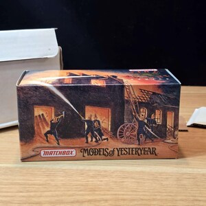 May include: A Matchbox Models of Yesteryear box features a vintage illustration of firefighters battling a blaze. The artwork depicts a burning building, firefighters with a hose, and a ladder. The box is brown with the Matchbox logo.