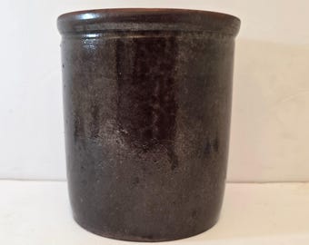 May include: A dark brown stoneware crock with a cylindrical shape. The antique jar has a slightly flared rim and a glossy finish. The vintage kitchenware is likely used for food storage or decoration.