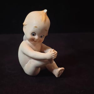 May include: A vintage bisque Kewpie doll figurine. The doll is seated with its knees bent and arms wrapped around its legs. The doll has a smiling expression, rosy cheeks, and a tuft of blonde hair. The figurine is on a black background.