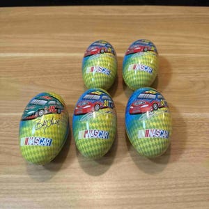 May include: Five NASCAR-themed egg-shaped containers. Each container features a race car design with the NASCAR logo. The containers have a yellow and green diamond pattern background. One container has the signature of Bobby Labonte.