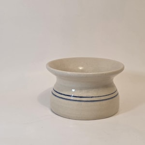 May include: A small, off-white ceramic container with a flared top and two horizontal blue stripes. The handmade pottery piece has a cylindrical base and a slightly textured surface, suitable for holding small items.