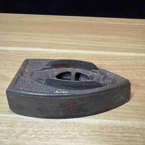 May include: A vintage, cast iron flat iron with a triangular shape. The iron is dark brown with a slightly rusted appearance, and features a handle rest and a central opening. The iron sits on a light brown wooden surface.