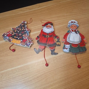 May include: Three vintage Christmas decorations: a Christmas tree, Santa Claus, and Mrs. Claus. The tree is decorated with ornaments and figures. Santa is in a red suit, and Mrs. Claus wears a red dress with a white apron. Each has a string and a red bead.