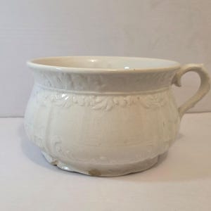 May include: Off-white ceramic chamber pot with a handle. The pot has a wide rim and a decorative floral pattern around the top and sides. The base is slightly raised.