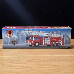May include: A red fire truck toy with a ladder, displayed on a box with a city skyline illustration. The box reads "5th in a Series" and "Authentic 1:25 Scale Model". The fire truck has silver details and black tires.