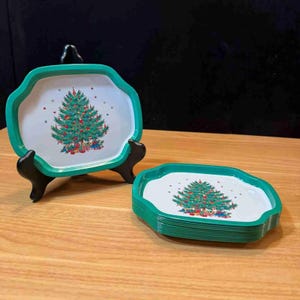 May include: A set of Christmas-themed serving trays. Each tray has a white interior with a green scalloped edge and a festive Christmas tree design. One tray is displayed on a black stand, while the others are stacked on a wooden surface.