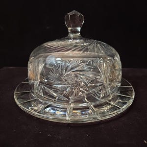 May include: A clear, cut-glass butter dish with a domed lid and a faceted knob. The dish features intricate starburst and geometric patterns, and sits on a round base. The glass reflects light, creating a sparkling effect.
