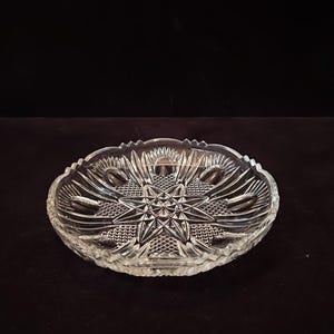 May include: Clear glass dish with a starburst design in the center. The dish has a scalloped edge and is made of clear glass. The starburst design is surrounded by a textured pattern. The dish is likely used for serving or display.