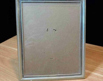May include: A rectangular silver-colored picture frame with a beaded border. The frame is empty, with a cardboard backing visible. The frame is sitting on a wooden surface.