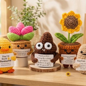 May include: A collection of five crocheted figures including a yellow duck, a pink flower in a pot, a brown poop shape, a yellow sunflower in a pot, and a beige potato, each holding a white sign with printed text and small yellow ducks.