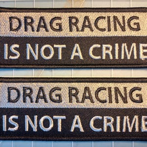 May include: Two black embroidered patches with the text "DRAG RACING IS NOT A CRIME" in white and silver thread. The text is arranged in two lines, with "DRAG RACING" above "IS NOT A CRIME".