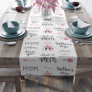 May include: White Mother's Day table runner with black and pink text. The runner features phrases like "Happy Mother's Day," "Best Mom Ever," and "Love You," along with heart and floral designs. The runner is set on a wooden table with place settings.