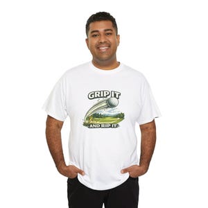 May include: White t-shirt with a golf-themed graphic. The design features a golf ball in motion, a golf course scene, and the text "GRIP IT AND RIP IT" in a bold font. The shirt is made of a soft material.