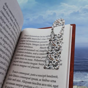 May include: A patterned bookmark with small illustrations of vehicles and figures sits in an open book. The bookmark has a white background with black and brown designs. The book has red edges and is open to a page of text.