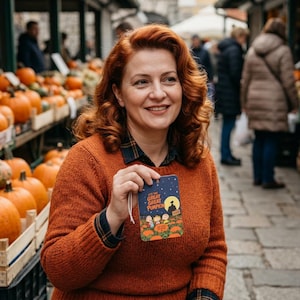 Pode incluir: Uma etiqueta retangular com um céu noturno azul-escuro, uma lua amarela e o texto 'IT'S THE GREAT GREAT PUMPKIN' sobre um campo de abóboras cor de laranja e personagens de desenhos animados, segurada perante muitas abóboras em caixotes.