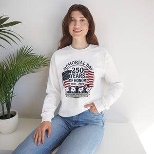 Op de afbeelding: Witte sweatshirt met lange mouwen met een Memorial Day-afbeelding. Het ontwerp bevat een Amerikaanse vlag, de tekst "Memorial Day 250 Years of Honor 1776-2026" en silhouetten van soldaten. Het sweatshirt wordt door een persoon gedragen.