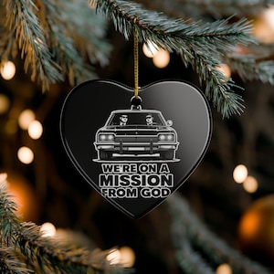 May include: Black heart-shaped Christmas decoration with a gold hanging string. The ornament displays a white illustration of a vintage car with two figures inside. The text "WE'RE ON A MISSION FROM GOD" is below the car.