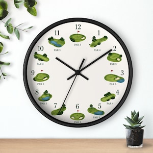 Puede incluir: Un reloj de pared redondo con marco negro y esfera blanca. La esfera presenta ilustraciones de hoyos de golf, cada uno con un valor de par. Las manecillas negras indican la hora. Una pequeña planta en maceta está sobre una superficie de madera.