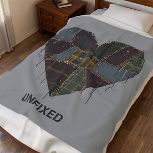 May include: A gray bedspread with a large heart design composed of patchwork fabric in shades of blue, green, and burgundy. The word "UNFIXED" is printed in black at the bottom. The heart is stitched with gold thread.