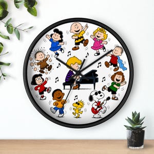 May include: A round wall clock with a black frame featuring a Peanuts character illustration. Schroeder plays a black piano in the center, surrounded by Charlie Brown, Lucy, Linus, Sally, Franklin, Peppermint Patty, Marcie, Snoopy, and Woodstock.