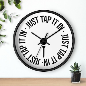 Puede incluir: Un reloj de pared redondo en blanco y negro con el texto "JUST TAP IT IN" que rodea la esfera del reloj. Las manecillas del reloj son negras y un gráfico de tee de golf está en el centro. El reloj está sobre una superficie de madera.