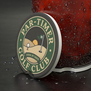 May include: A round golf club coaster with the text "PAR-TIMER GOLF CLUB" in green and white. The coaster features a golf course graphic with a golf ball, flag, and sun. The coaster is resting next to a glass.