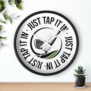 Könnte beinhalten: Eine runde, schwarze Wanduhr mit dem Text "JUST TAP IT IN" in einem kreisförmigen Muster. Das Zifferblatt zeigt einen Golfschläger, einen Golfball und grüne Akzente. Die Zeiger sind weiß.