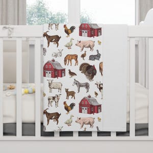 May include: A white baby blanket featuring a repeating pattern of farm animals and red barns. The illustrated animals include goats, chickens, pigs, ducks, sheep, donkeys, and cows. The blanket is draped over a white crib.