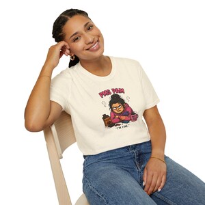Può includere: T-shirt color crema con un'illustrazione a cartoni animati di una donna arrabbiata, la scritta "PMS PAM" e "I'M FINE." La grafica include una tazza di caffè e una bottiglia. La maglietta è indossata da una persona.