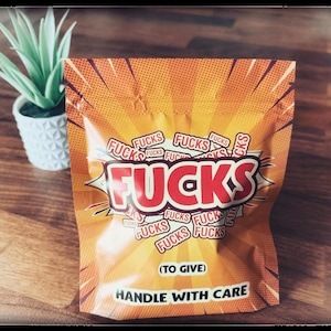 May include: An orange and yellow package with the word "FUCKS" in large red letters. The package has the words "FUCKS" repeated in smaller text and the phrases "(TO GIVE)" and "HANDLE WITH CARE". A small potted plant is in the background.
