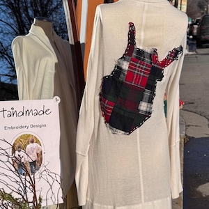 May include: Cream-colored long-sleeve sweater with a turtleneck. The back features a patchwork design in red, green, and black plaid. The sweater is displayed on a mannequin outdoors, with a mirror and a sign that says "Handmade Embroidery Designs" in the background.