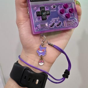 May include: A clear purple handheld gaming device case with a matching purple lanyard and a Gengar charm. The case has a button layout and a black wrist strap.