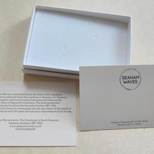K&ouml;nnte beinhalten: Eine wei&szlig;e Box mit einer wei&szlig;en Karte darin. Die Karte enth&auml;lt den Text "Seaham Waves" und eine Beschreibung des handgefertigten Schmucks des Unternehmens, der aus Meerglas hergestellt wird, das an der K&uuml;ste von Seaham, County Durham, England, gesammelt wird. Der Text enth&auml;lt auch die Website und die Telefonnummer des Unternehmens.
