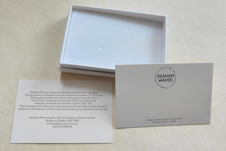 K&ouml;nnte beinhalten: Eine wei&szlig;e Box mit einer wei&szlig;en Karte darin. Die Karte enth&auml;lt den Text "Seaham Waves" und eine Beschreibung des handgefertigten Schmucks des Unternehmens, der aus Meerglas hergestellt wird, das an der K&uuml;ste von Seaham, County Durham, England, gesammelt wird. Der Text enth&auml;lt auch die Website und die Telefonnummer des Unternehmens.