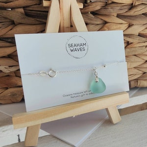 May include: A silver chain bracelet with a light blue sea glass charm. The bracelet is displayed on a wooden easel with a white card that reads "Seaham Waves" and "Oceans treasure left on Nature's gift to adore."