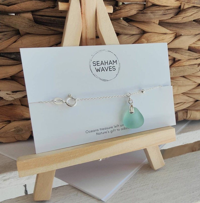 May include: A silver chain bracelet with a light blue sea glass charm. The bracelet is displayed on a wooden easel with a white card that reads "Seaham Waves" and "Oceans treasure left on Nature's gift to adore."