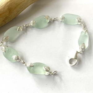 K&ouml;nnte beinhalten: Ein silbernes Armband mit einer Kette und hellgr&uuml;nen Meerglassperlen. Jede Perle ist mit einem silbernen Anh&auml;nger verziert.