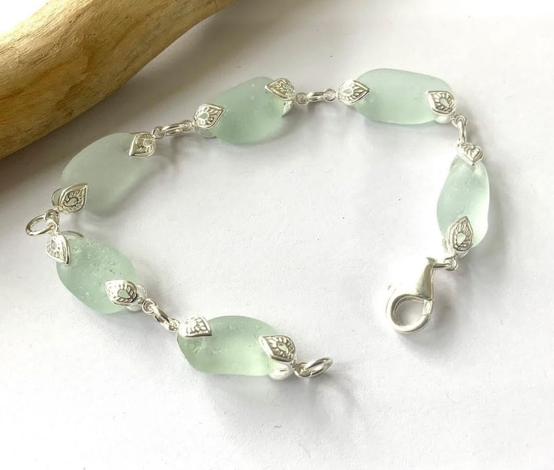 K&ouml;nnte beinhalten: Ein silbernes Armband mit einer Kette und hellgr&uuml;nen Meerglassperlen. Jede Perle ist mit einem silbernen Anh&auml;nger verziert.