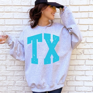 May include: A light gray sweatshirt with the turquoise letters "TX" printed on the front. The sweatshirt has long sleeves and a crew neck. The person in the image is wearing a black and white baseball cap.